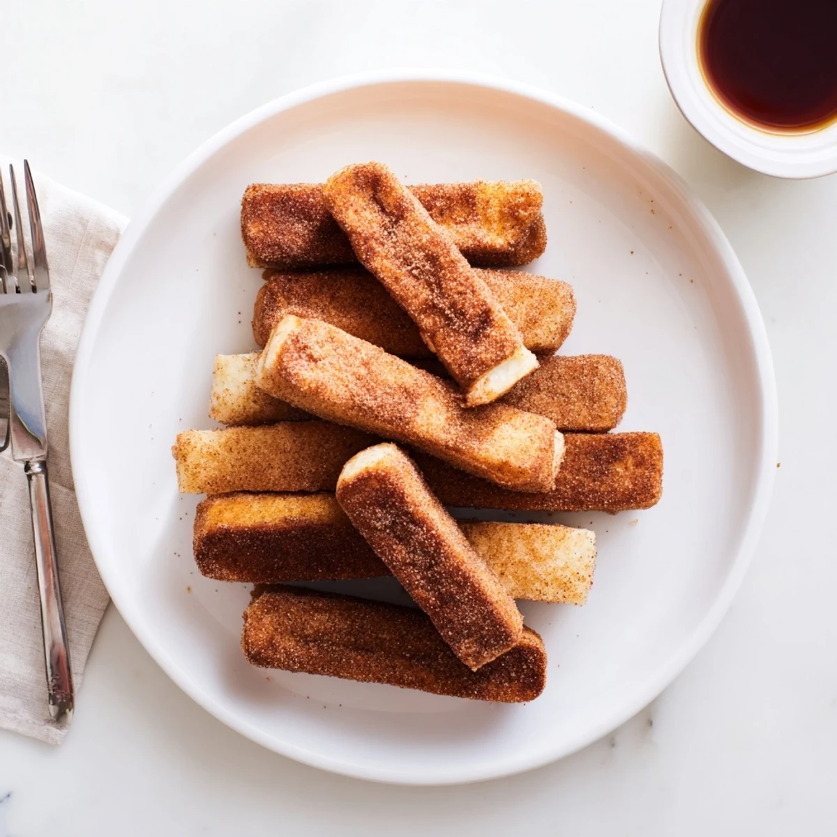 Golden cinnamon French toast sticks arranged on a white plate with maple syrup pooling beside them.