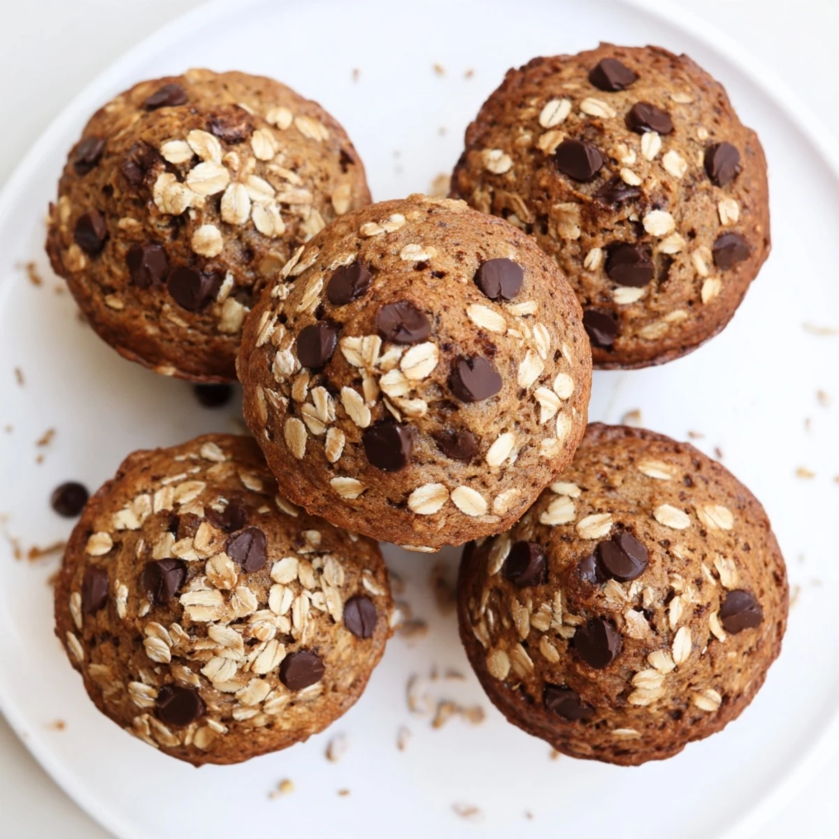 Golden banana chocolate chip muffins with oats stacked on a wooden serving board, featuring melted chocolate spots and a crumb-topped texture.